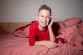 A cute boy in red pajamas is lying in bed under a blanket and talking on the phone Royalty Free Stock Photo