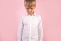 A cute boy stands next to pink wall, whites hirt, hands in pockets, bowing his head down