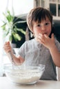 A cute boy, a toddler of Caucasian appearance, cooks in the kitchen and tastes the dough. Vertical photo. Ideal for illustrating