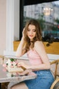 Cute brown-haired young girl sitting in a cafe outdoor, rattan chairs, reading an open book Royalty Free Stock Photo