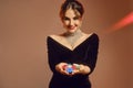 Brunette female in black dress and shiny jewelry. Smiling, showing handful of colorful chips, posing on brown background