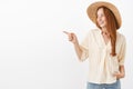 Cute carefree and stylish female redhead traveler in trendy straw hat and yellow summer blouse pointing and gazing left