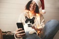 Cute cat looking at phone screen with funny emotions and sitting on happy girl legs in christmas lights. Young hipster woman in Royalty Free Stock Photo