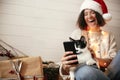 Cute cat looking at phone screen with funny emotions and sitting on happy girl legs in christmas lights. Young hipster woman in Royalty Free Stock Photo