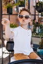 Cute cauasian boy in white tee shirt and sunglasses sitting at the table in loft style cafe and pointing somewhere.