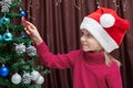 A cute Caucasian blonde girl in a red sweater and a Christmas cap hangs candy on a decorated Christmas tree Royalty Free Stock Photo