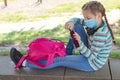 A cute Caucasian girl in a mask sits in the schoolyard with a backpack and a phone in her hands. Gadgets and kids. Rest from study