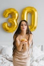 Cute Caucasian girl in a nightgown, who is congratulated on her birthday in bed, blows out a candle on a cake and makes a wish for