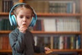 Cute Caucasian little girl in big white headphones listens music in the interior against the background of bookshelves Royalty Free Stock Photo