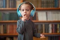 Cute Caucasian little girl in big white headphones listens music in the interior against the background of bookshelves Royalty Free Stock Photo