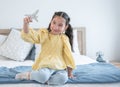 Cute Caucasian little girl with pony tails hair sitting on bed at home and playing with airplane model toy, holding plane to fly Royalty Free Stock Photo