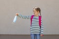 A cute Caucasian schoolgirl with a knapsack behind her back holds a medical mask on her outstretched hand and makes a face.