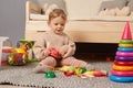 Cute charming baby girl wearing warm beige jumper sitting on floor near sofa and playing with toys alone at home, early Royalty Free Stock Photo