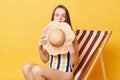 Cute charming surprised woman wearing striped swimming suit sitting on deck chair isolated over yellow background covering half of Royalty Free Stock Photo