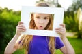 Cute cheerful little girl holding white picture frame in front of her face