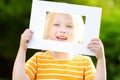 Cute cheerful little girl holding white picture frame in front of her face