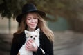 Cute cheerful slim caucasian girl blonde with long hair in a black coat and a black hat in in the autumn day with her white small Royalty Free Stock Photo