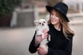 Cute cheerful slim caucasian girl blonde with long hair in a black coat and a black hat in in the autumn day with her white small