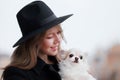 Cute cheerful slim caucasian girl blonde with long hair in a black coat and a black hat in in the autumn day with her white small