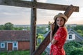 Cute cheerful teen girl in red dress and hat holds on to a wooden support and looking at the camera, landscape on the