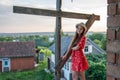 Cute cheerful teen girl in red dress and hat holds on to a wooden support and looking at the camera, landscape on the