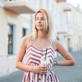 Cute cheerful young woman blonde in a trendy pink striped dress is standing in the city and drinking a milky sweet cocktail. Royalty Free Stock Photo