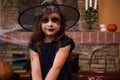 Cute child girl enchantress, in wizard& x27;s hat, sits against a cobweb-covered fireplace with spell books, smiles at Royalty Free Stock Photo