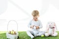Child holding white box while sitting on green grass near toy rabbit and straw basket with Easter eggs isolated on white Royalty Free Stock Photo