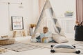 Cute child reading a book, laying on the pillows in stylish scandinavian tent in playroom