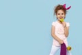 Cute child in stylish outfit with lollipop looking at camera while posing with violet longboard on blue background. Royalty Free Stock Photo