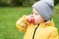 A cute child in a yellow raincoat against a background of green greedily bites an apple and looks to the side. Country life on a