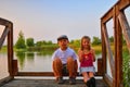 Cute children, little boy and girl are sitting on pier in the summer evening. Two siblings. Golden hour by the lakeside. Happy chi