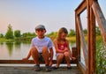 Cute children, little boy and girl are sitting on pier in the summer evening. Two siblings. Golden hour by the lakeside. Happy chi Royalty Free Stock Photo