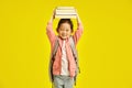 Cute chinese schoolgirl holding books on head over yellow  background. First grader kid girl preparing to the Royalty Free Stock Photo