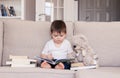 Cute clever little baby boy keen about reading book sitting on sofa with teddy bear toy and pile of books at home. Royalty Free Stock Photo