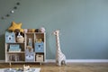 Cute composition of stylish scandi child room interior design with armchair, wooden shelf, plush giraffe, toys.