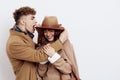 a cute couple is standing on a white background, wearing a coat. The man gently touches the ends of the hat, absurdly