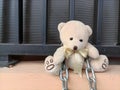 Cute cream-colored teddy bear with a ribbon necklace sits tied on chain and black iron fence outdoors