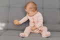 Cute curious female child wearing striped sleeper sitting on gray couch with her toy in hand, playing at home, having fun, happy Royalty Free Stock Photo