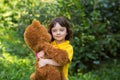 Cute curly girl in a yellow T-shirt smiles and holds a large brown teddy teddy bear. greenery around Royalty Free Stock Photo