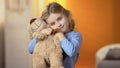 Cute curly-haired blond girl hugging teddy bear and smiling at camera, happiness Royalty Free Stock Photo