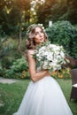 A cute curly woman in a white wedding dress with a wedding bouquet and wreath in her hair standing back to the camera in nature.