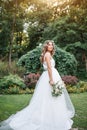 A cute curly woman in a white wedding dress with a wedding bouquet and wreath in her hair standing back to the camera in nature.