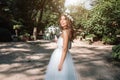 A cute curly woman in a white wedding dress with a wedding bouquet and wreath in her hair standing back to the camera in nature. C