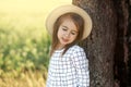 A cute cute little girl in a hat and a checkered white dress is standing by the trunk of a large tree. Royalty Free Stock Photo
