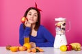 Cute dark haired thoughtful girl with red hair band and blue shirt holds apple in hand. Woman makes fruit smoothie. Lady uses