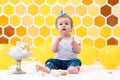 A cute dirty toddler is sitting next to a broken cake. In the background is a pattern of yellow honeycombs and balloons