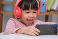 Cute elementary school girl wearing headphones holding a smartphone. Happy Asian kid studying online on smartphone or Royalty Free Stock Photo