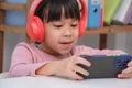 Cute elementary school girl wearing headphones holding a smartphone. Happy Asian kid studying online on smartphone or Royalty Free Stock Photo