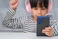 Cute elementary school girl wearing headphones holding a smartphone. Happy Asian girl studying online on smartphone or Royalty Free Stock Photo